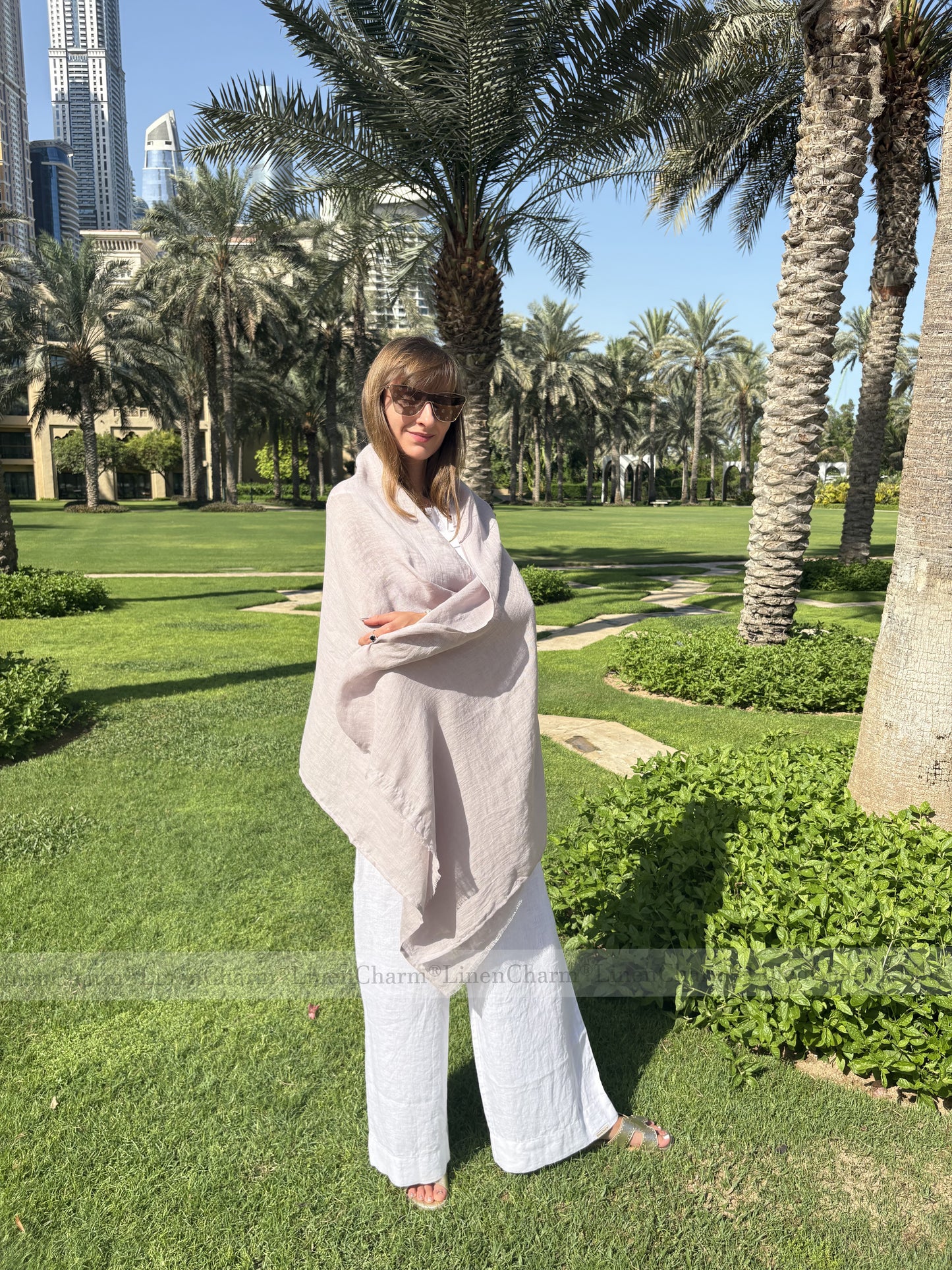 Woman in a light-colored outfit standing in a park with palm trees and modern buildings in the background.