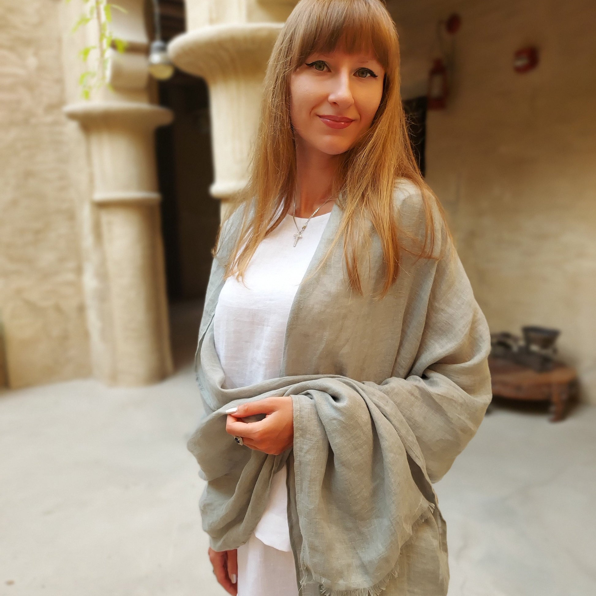 A woman standing outdoors wearing a soft green linen scarf with tassels on the ends, paired with a white top.