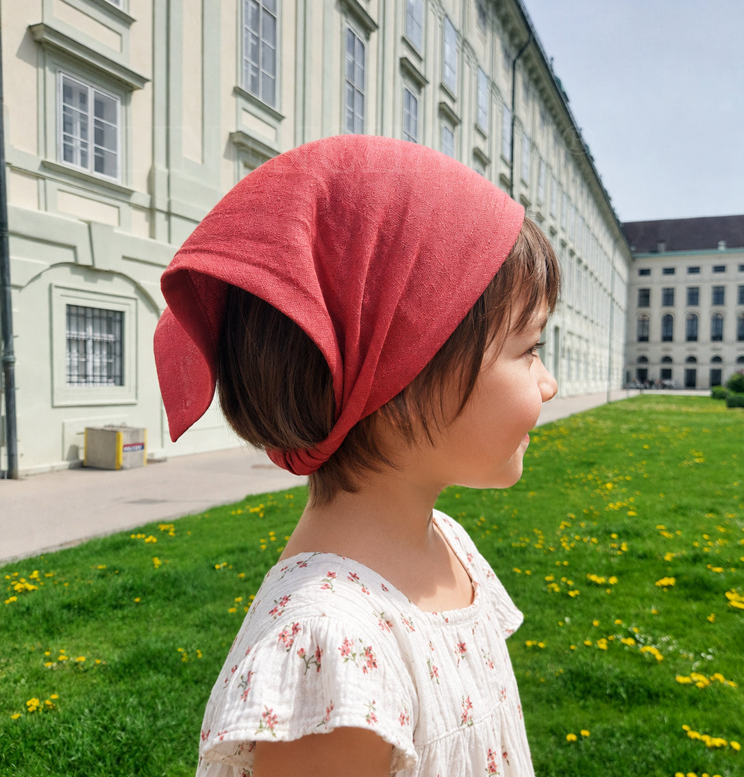Child wearing a red headscarf in front of a large building with green grass and yellow flowers.