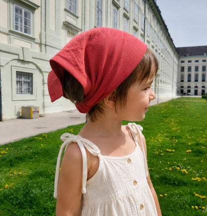 Child wearing a red headscarf and white dress in front of a building with green grass and yellow flowers.