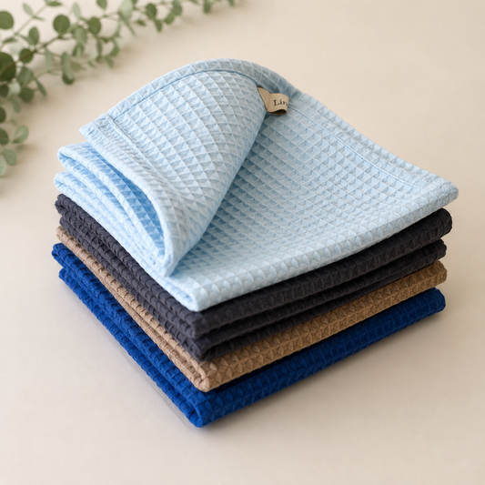 A stack of textured waffle-weave cloths or baby wipes in light blue, dark gray, tan, and royal blue, arranged on a light beige surface with a small leafy sprig in the corner.