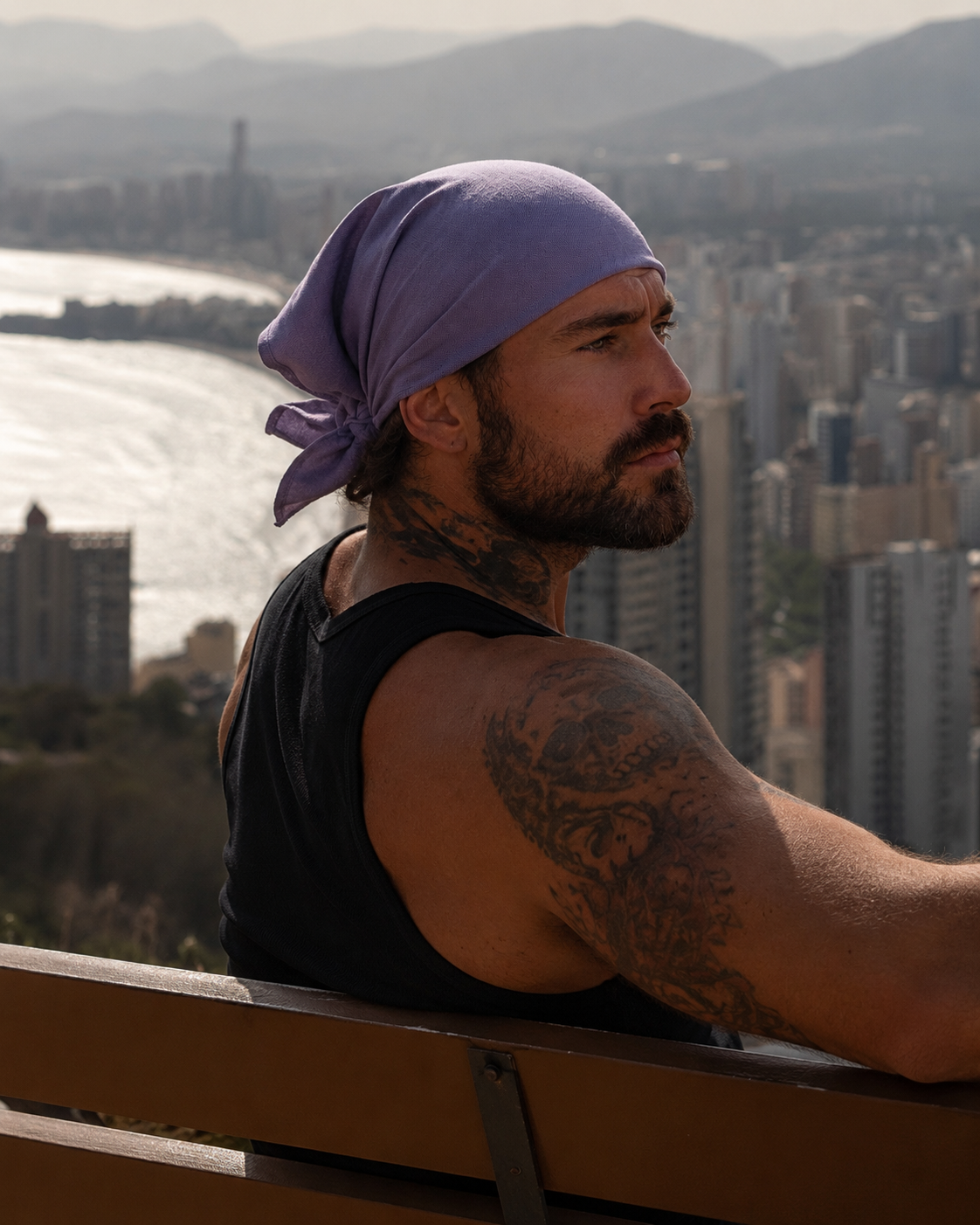 A man with a beard and tattoos wearing a purple bandana and a black tank top, sitting on a wooden bench with a city and river in the background.