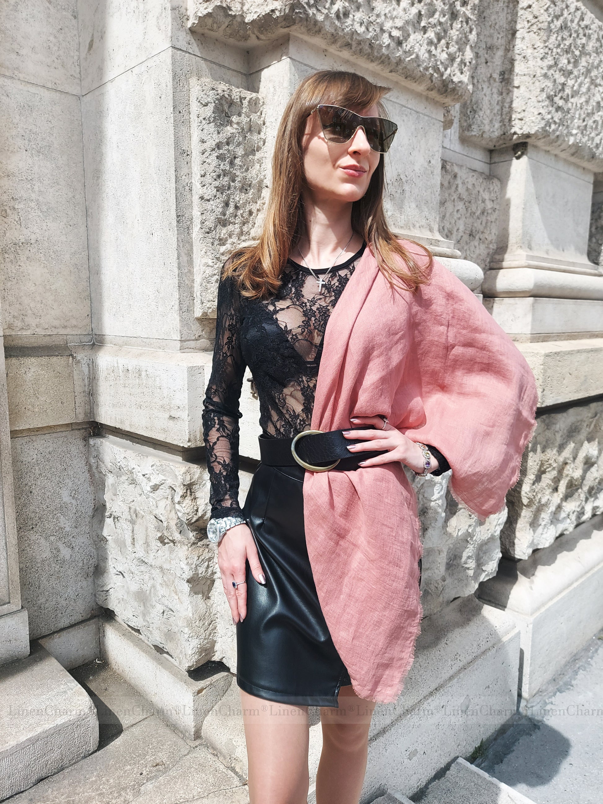 Woman wearing a black lace top and leather skirt with a pink linen scarf, standing against a stone wall.