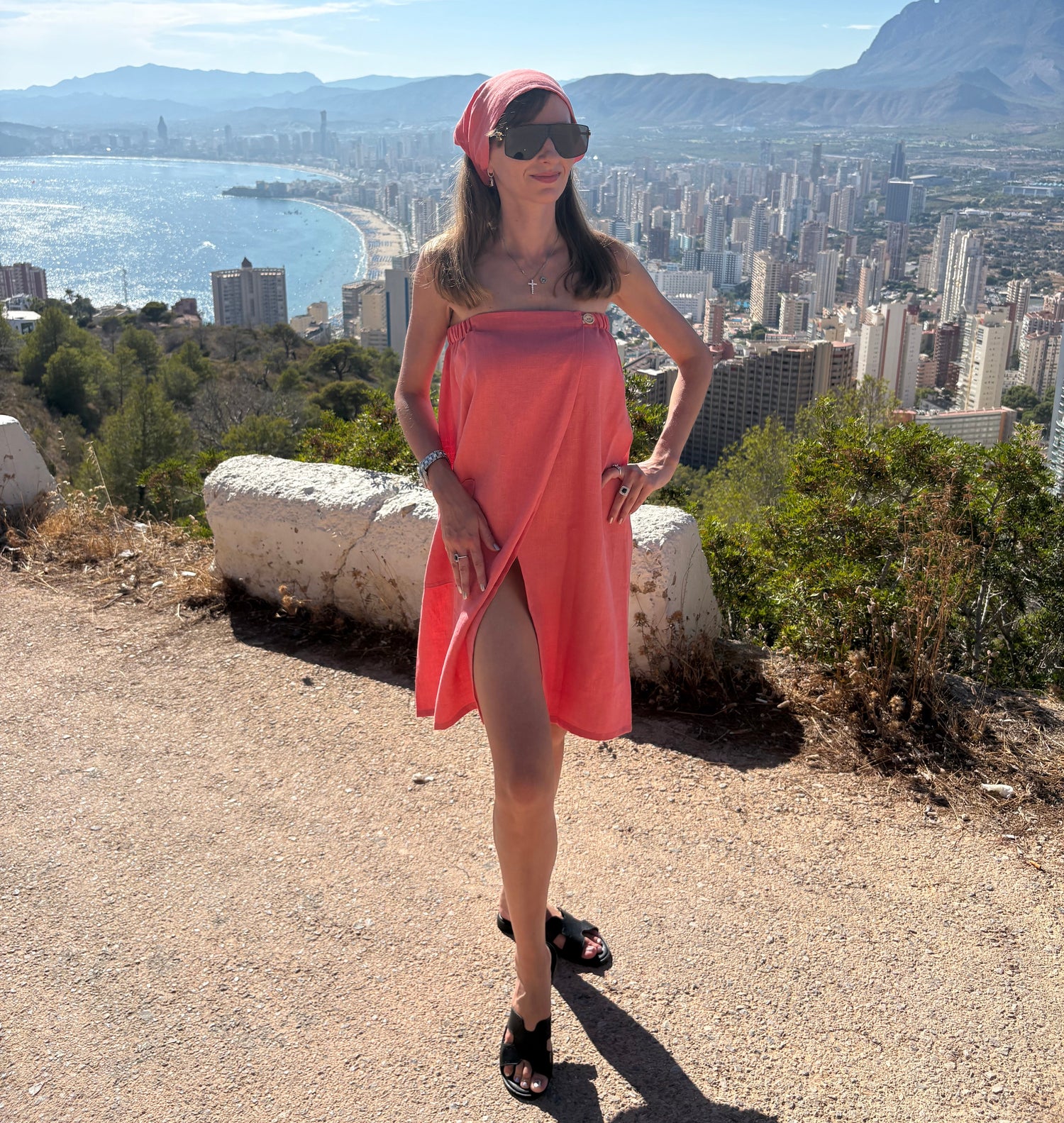 A woman wearing a bright coral-pink headscarf and matching strapless dress stands on a sunny outdoor overlook with a panoramic city and beach view in the background.